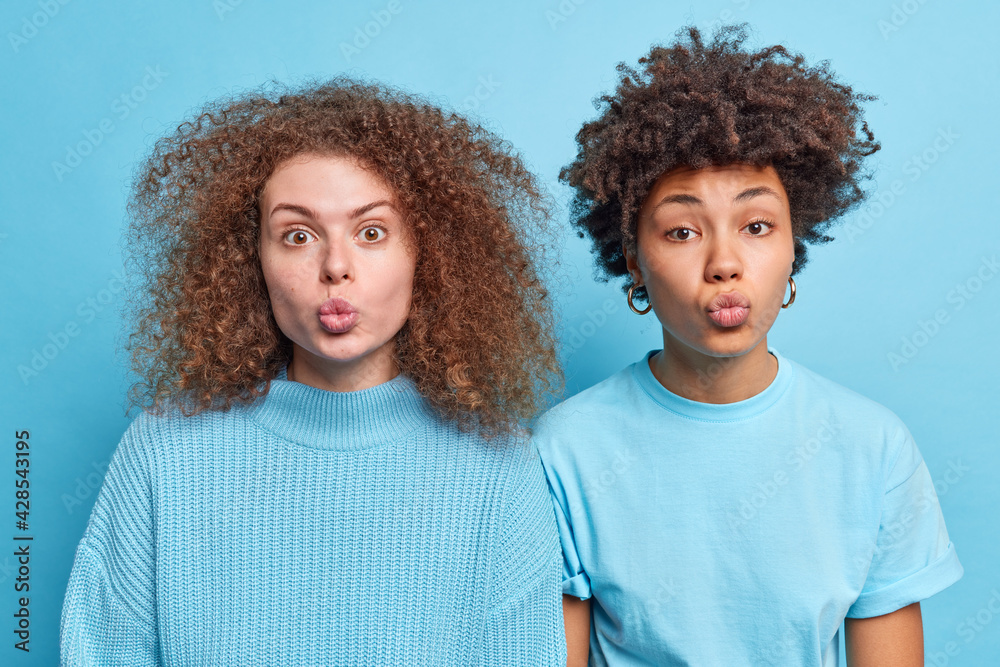 Wall mural Two diverse young women stand next to each other keep lips folded want to kiss someone have curly bushy hair wear casual clothes isolated over blue background. Human face expressions concept