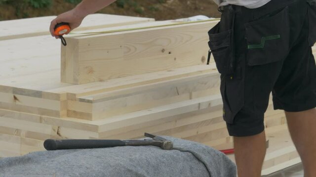 CLOSE UP: Unrecognizable contractor in black cargo shorts measures a CLT beam and marks it with a pencil while working at construction site. Builder measuring and marking a glued-laminated timber beam