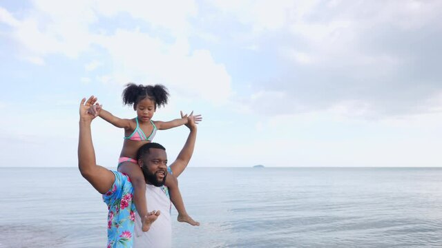 Happy mixed race family on summer vacation. African father carrying little daughter and walking on the beach. Black dad and cute child girl kid enjoy and having fun outdoor activity playing together