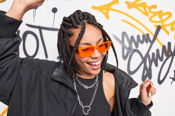 Glad teenager with trendy hairstyle dances carefree keeps arms raised enjoys spending time in public place wears orange sunglasses black jacket poses against drawn graffiti wall. Street art concept