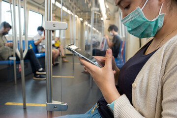 Young Asian Japanese woman wearing face mask and using smart phone in BTS subway train Bangkok...