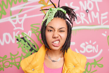 Teenage lifestyle concept. Cheeky hipster girl with combed dreadlocks clenches teeth smirks face looks self confident at camera spends free time at street poses over graffiti wall background - obrazy, fototapety, plakaty