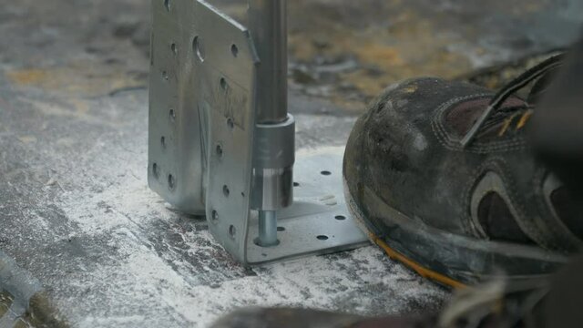 SLOW MOTION, CLOSE UP, DOF: Contractor fastens a stainless steel wall carrier into the ground. Unrecognizable builder uses a power drill to fasten a bolt into bitumen covered concrete foundation.