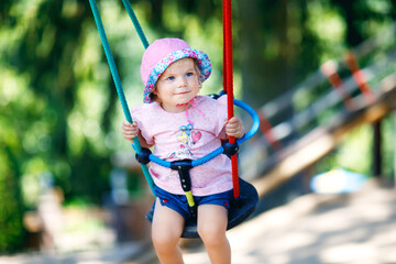 Cute adorable toddler girl swinging on outdoor playground. Happy smiling baby child sitting in...