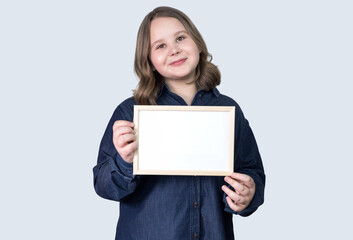 A teenage girl stands in a blue shirt and holds a frame on a light background. She's happy. She's Caucasian. Copy space. Soft focus. Mock up.