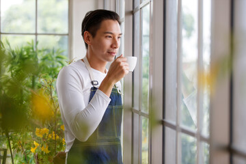 Handsome man short black hair wearing t-shirt jean apron holding coffee cup dimple smiling standing. Photo blur selective focus his warm face. Indoor with sunlight and air filtering plants.