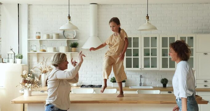 Little artistic girl perform dances on dining table having fun together with young carefree mom and elderly 60s granny in cozy kitchen on weekend. Family home hobby, active quarantine with kid concept
