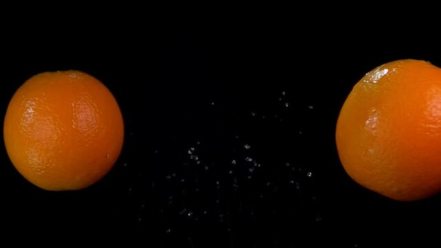 Large juicy oranges are flying and colliding with each other rising drops of water on the black background in slow motion