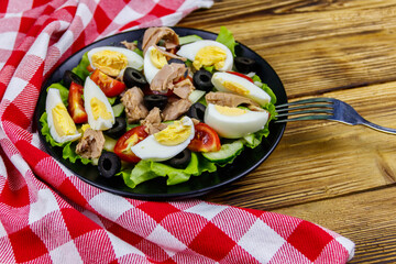 Tasty tuna salad with lettuce, black olives, eggs and fresh vegetables on wooden table