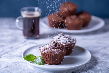 Chocolate muffins with mint on a white plate, strewed with icing sugar. Homemade baking. In the background is a cup of coffee and a plate with muffins. Marble table and blue background. Close up.