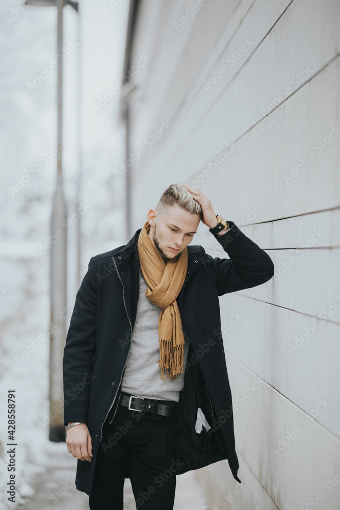 Wall mural Handsome man in a black winter outfit with a brown scarf leaning against a wall with his arm