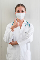 Happy Caucasian young female doctor wearing protective facemask and white hospital gown gives a thumbs up and look at camera, vertical half-length portrait on white background. 