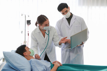 Fototapeta premium Caucasian female and Asian male doctors wearing face mask and hospital white gown discuss Asian male patient’s online medical report from laptop at hospital ward. Medical and healthcare concept.