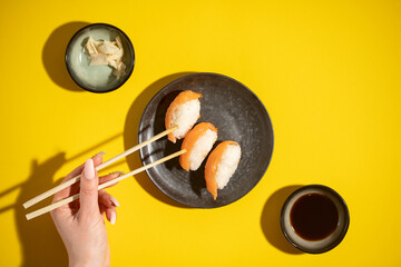 Sushi nigiri with salmon on black plate on yellow background, with soja sauce and ginger and female hand with chopsticks, top view flatlay