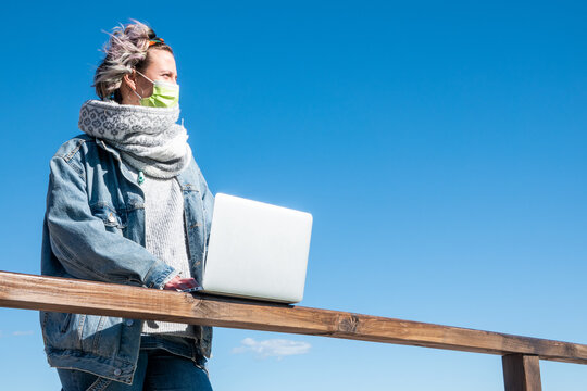 Spanish Woman Holding A Laptop While Standing Beside Wooden Railings Against Blue Sky Background