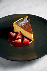 Piece of delicate curd casserole with creamy and berry sauce and strawberry slices in black plate on white marble background. Cheesecake