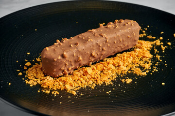 Dessert bar of ice cream with brownie flavor, with milk chocolate glaze with nuts and decoration of crunch on a black plate on a white marble background.
