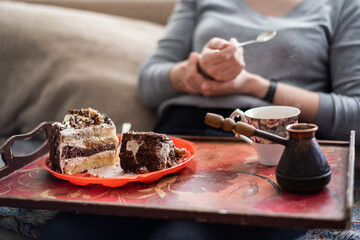 A woman is having breakfast at home, eating a cake with coffee. A piece of cake and a coffee turk on a tray.