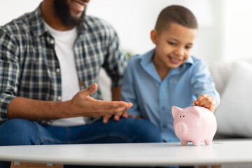 Father And Son Putting Personal Savings In Piggybank Indoors, Cropped