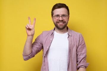 Joyful man in glasses and casual clothes shows the symbol of victory and looks at the camera smiling. Digit with two fingers. 