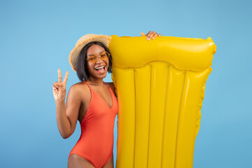 Cheerful black lady in swimsuit holding yellow lilo and showing peace gesture on blue studio...