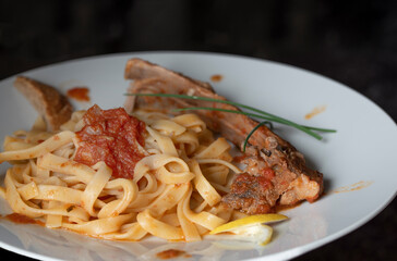 A lunch with lamb ribs on ribbon noodles with tomato sauce in front of a dark background
