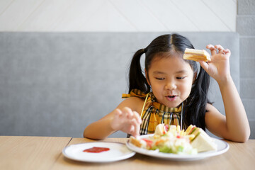 Asian child hungry or kid girl smile to enjoy happy eating bread or sandwich with ketchup and vegetable salad on dish and table at school or home kitchen and restaurant for breakfast food or snack