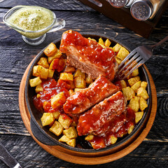 baked meat with fried potato cubes and tomato sauce in a saucepan on a black textured wood background