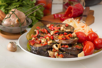 Pickled vegetables - eggplants in soy sauce with pepper and garlic, cucumbers, tomatoes, cabbage in a white plate against the background of glass jars with homemade pickles and herbs. Close-up