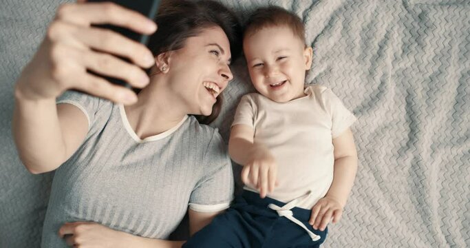 Laughing woman and baby boy making a selfie or video call in a bed. Concept of technology, new generation, family, connection, parenthood. Top view, slow motion.