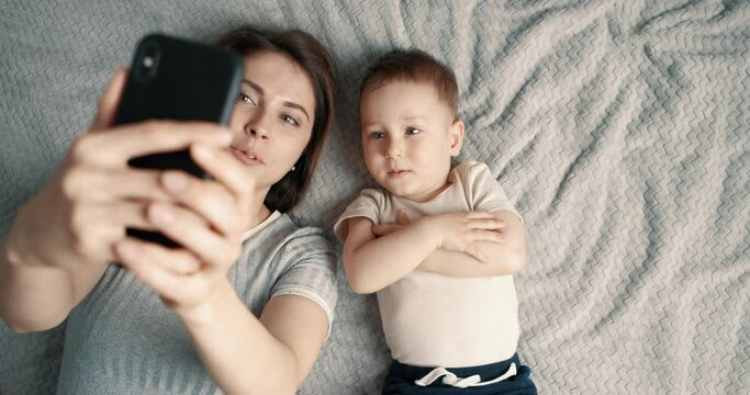 Mom and son take selfies using mobile phone. A woman and a small baby boy lie on bed and look at smartphone camera. Video call. Top view, slow motion