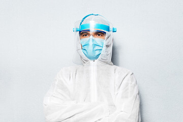 Fototapeta na wymiar Portrait of doctor man with crossed arms, wearing PPE suit against coronavirus and covid-19, on the background of white textured wall.