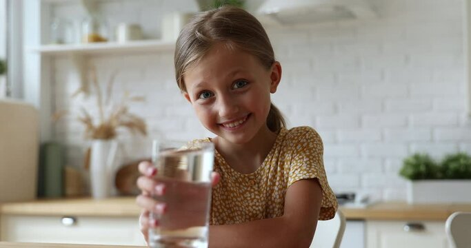 Beautiful caucasian little girl extend her hand to camera with glass offers still or mineral water. Good healthy habit, morning routine, body refreshment, daily aqua intake for stay hydrated concept