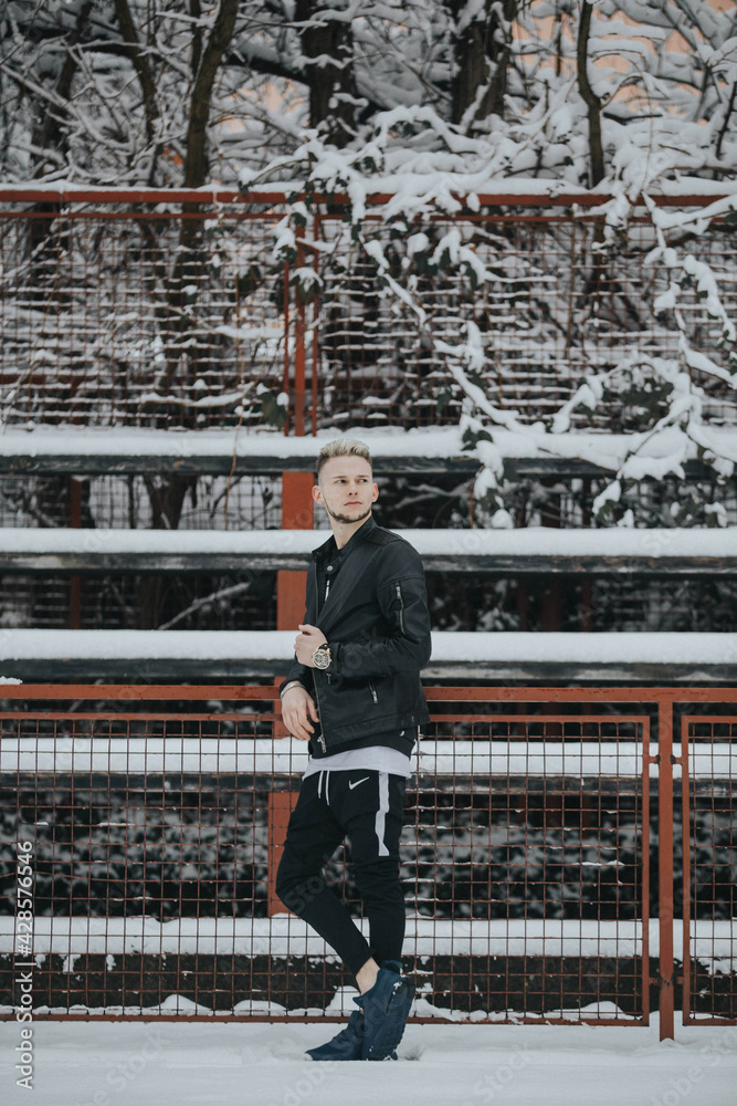 Wall mural Handsome young man in a black winter street style outfit leaning on a metal fence in a snowy park