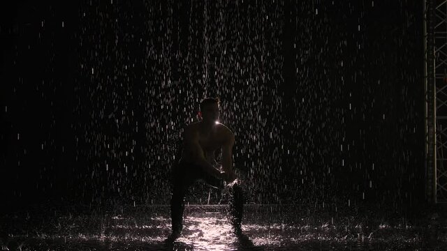 Silhouette of wet male dancer performing modern ballet dance in rain and splashing water. Male sensual modern choreography on black background in center of light beam. Slow motion.