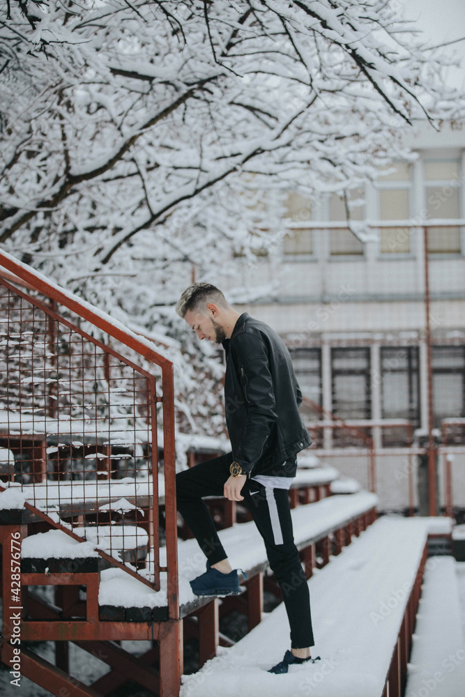 Wall mural Handsome young man in a black winter street style outfit climbing the snowy park stairs looking down