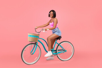 Summer mode. Pretty young black lady in trendy outfit riding bicycle on pink studio background, full length