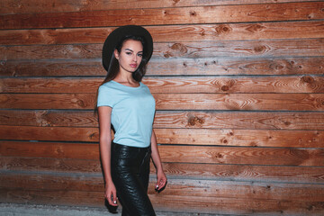 Hipster girl in hat, her hair straight dressed in leather pants and t shirt near the wooden wall,...