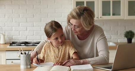 Loving 60s old grandmother helps to small schoolgirl granddaughter with homework girl writing on copybook, sit together in kitchen using laptop studying school subject. Homeschooling, tutoring concept - Powered by Adobe