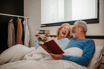 Photo of senior couple smiling in bed at home while reading a book.
