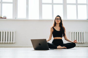 Beautiful brunette fitness woman meditate in front laptop, doing yoga indoors at home. Staying fit and healthy