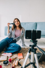 Attractive caucasian young woman video blogger showing beauty products via her blog on social media, holding makeup brushes looking at  smartphone camera fixed on tripod.