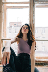 Attractive stylish caucasian woman with long brunette hair sitting on windowsill in living room.