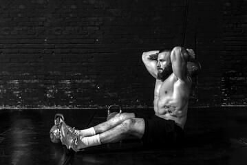 Young active sweaty strong muscular fit man with big muscles sitting on the floor of the gym and doing triceps workout with the heavy kettlebell as hardcore cross training real people exercising