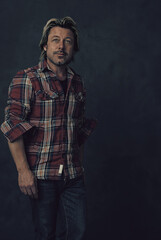 Middle-aged blond man with stubble in a lumberjack shirt and jeans standing in front of a dark wall.