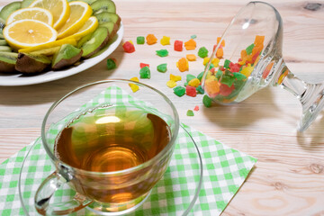 Glass cup of tea, multicolored candied fruits, a plate with lemon and kiwi slices on a wooden background with white and green napkins close-up