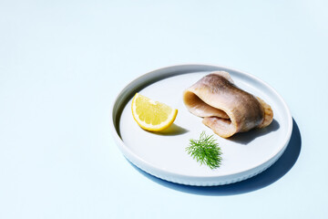Herring fillet rolled up with lemon slice and dill on the plate on light blue background. Minimal food concept. Classic marinated herring rolls.