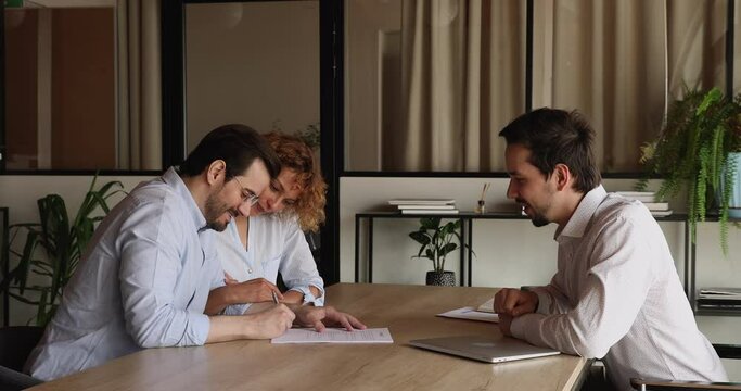 Side view smiling young family couple signing contract with professional hispanic realtor, celebrating making deal or purchasing first own apartment, taking keys from dwelling at meeting in office.