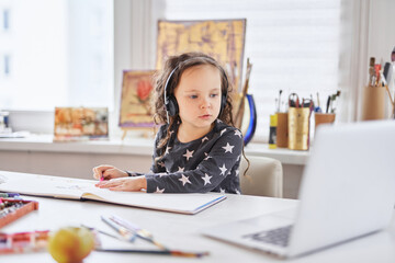 Online art classes at home during coronavirus lockdown or covid19 quarantine concept. Cute small kid girl in headphones sitting at a table using a laptop computer for education or homework doing. 