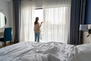 Fototapeta na wymiar Young asian woman holding cup of coffee and opening curtains in the morning and looking through the window at home
